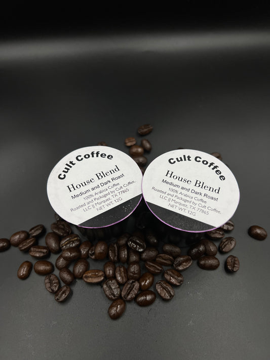 Two Cult Coffee single-serve pods labeled 'House Blend Medium and Dark Roast' are placed on a dark surface surrounded by scattered coffee beans. The labels indicate they are 100% Arabica coffee