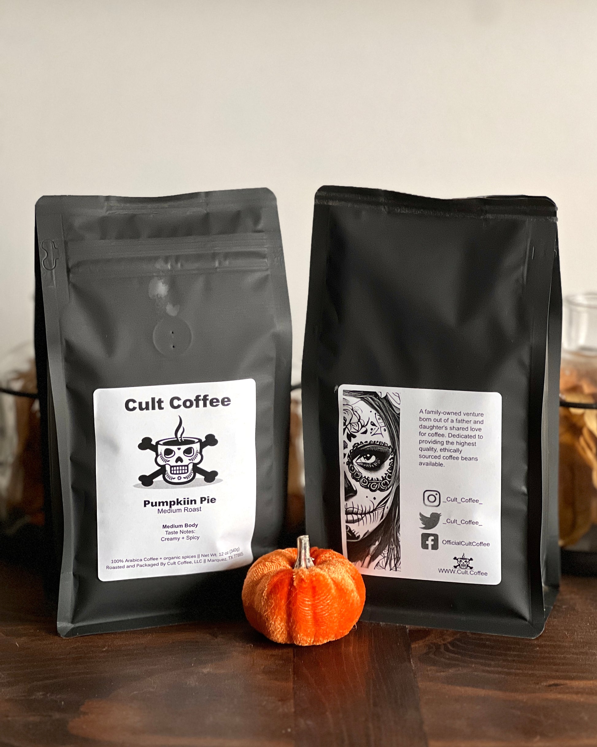 Two bags of Cult Coffee on a wooden counter; the left bag labeled 'Pumpkin Pie Medium Roast' with a skull and chef's hat graphic, and the right bag with a Day of the Dead-style face illustration. A small pumpkin sits in front. The background is minimal with a shelf and jars.